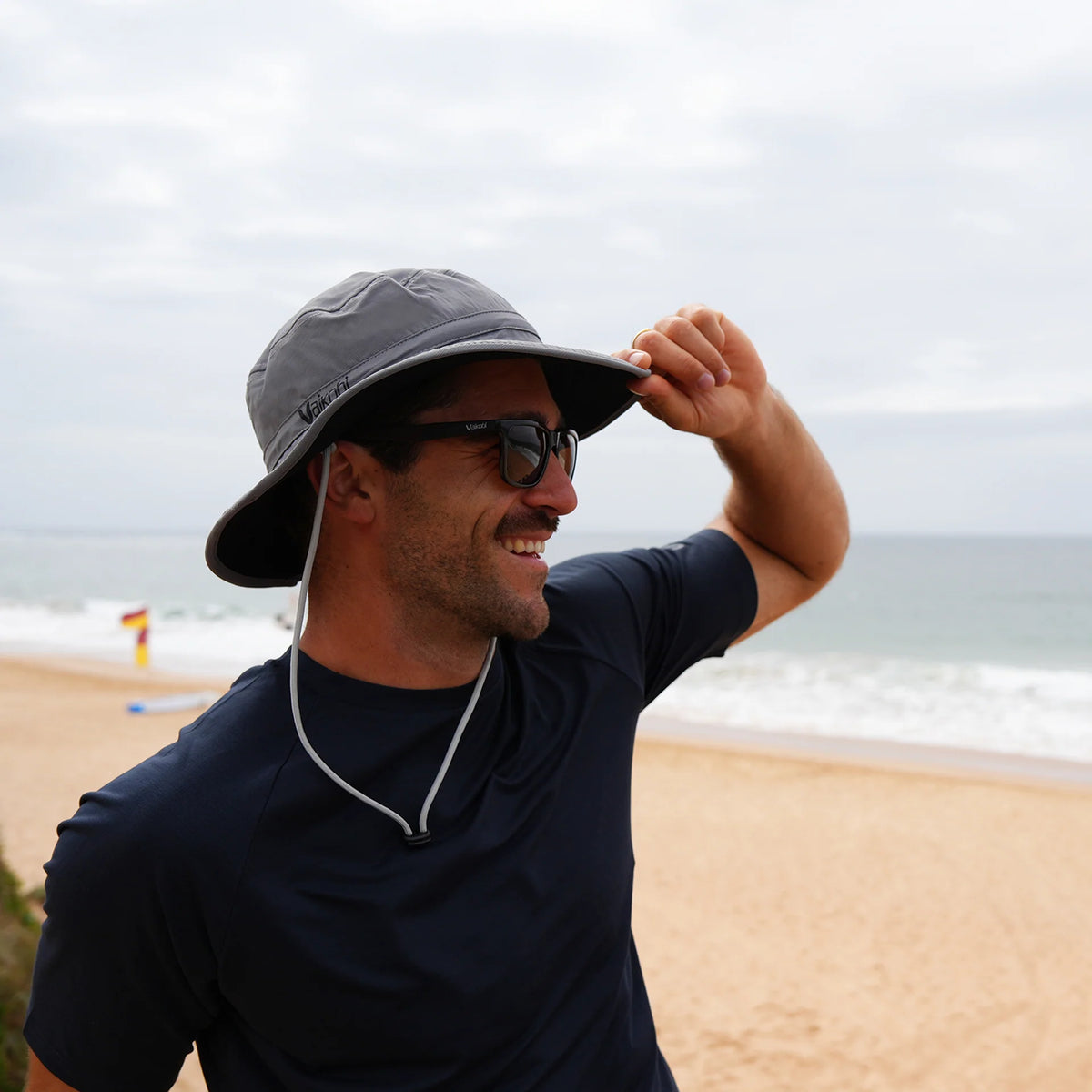 Man smiling and wearing Vaikobi UV Ocean Surf Hat in grey at the beach